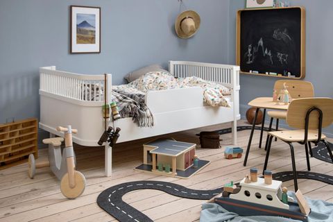 Kinderzimmer mit hellblauer Wandfarbe, weißem Juniorbett, Kreidetafel, Dreirad und auf dem Boden verteilten Spielsachen