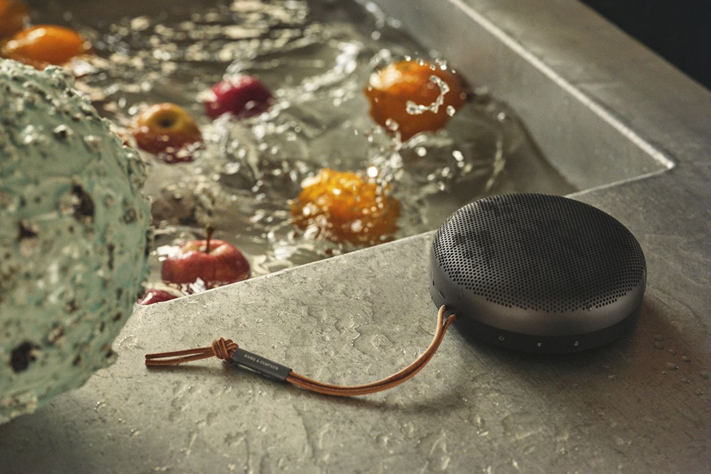Gefülltes Wasserbecken mit Obst drinnen, daneben: der mit Wasser bespritzte Bluetooth-Lautsprecher "Beosound A1" von Bang&Olufs…