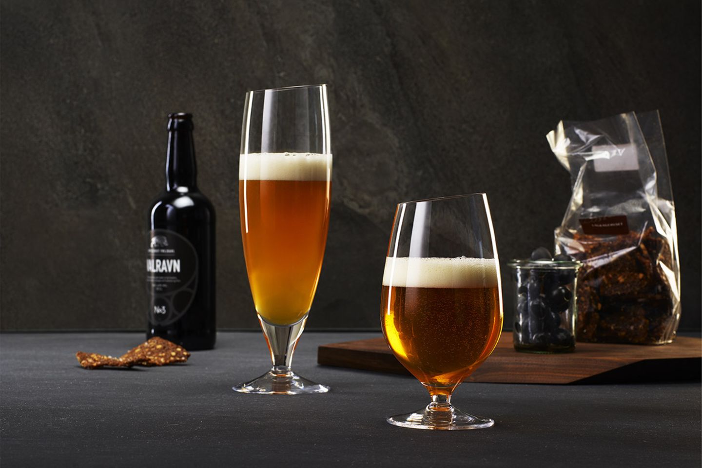 Zwei unterschiedlich große Biergläser, im Hintergrund stehen Oliven im Glas und Schokolade auf einem Holzbrett