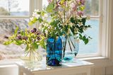 Blaue und durchsichtige Glasvasen mit Blumensträußen vor weißem Fensterrahmen