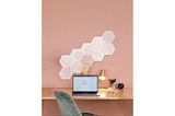 Weiß leuchtende Lichtpaneele in Hexagonform an pinker Wand vor einem Schreibtisch mit Laptop und Schreibtischlampe