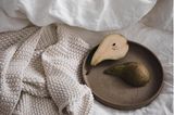 Weiße Bettwäsche, helle Strickdecke und ein dunkler Keramikteller mit Birne