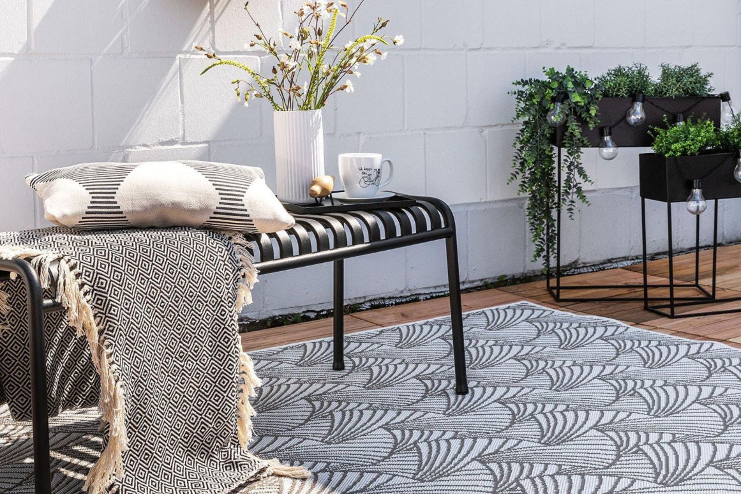Grauer Outdoor-Teppich auf Holzterrasse mit schwarzer Metallbank und Textilien