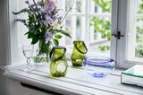 Fensterbrett mit zwei grünen Windlichtern, einer hellblauen Glasschale, einem Blumenstrauß und einem Glas Wasser