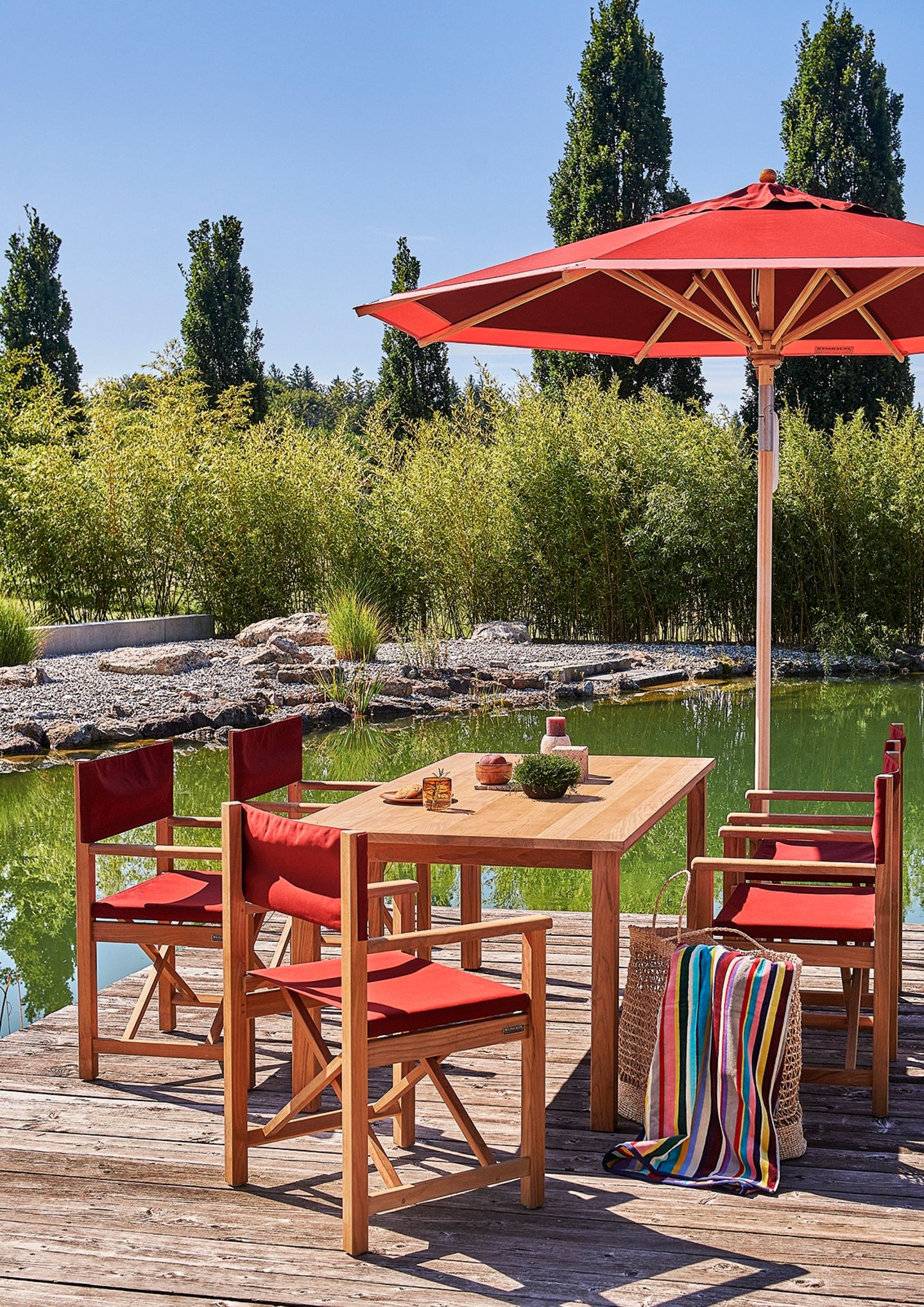 Sonnenschirm und Sitzgruppe in Rot und Holz auf einem Steg vor einem Teich