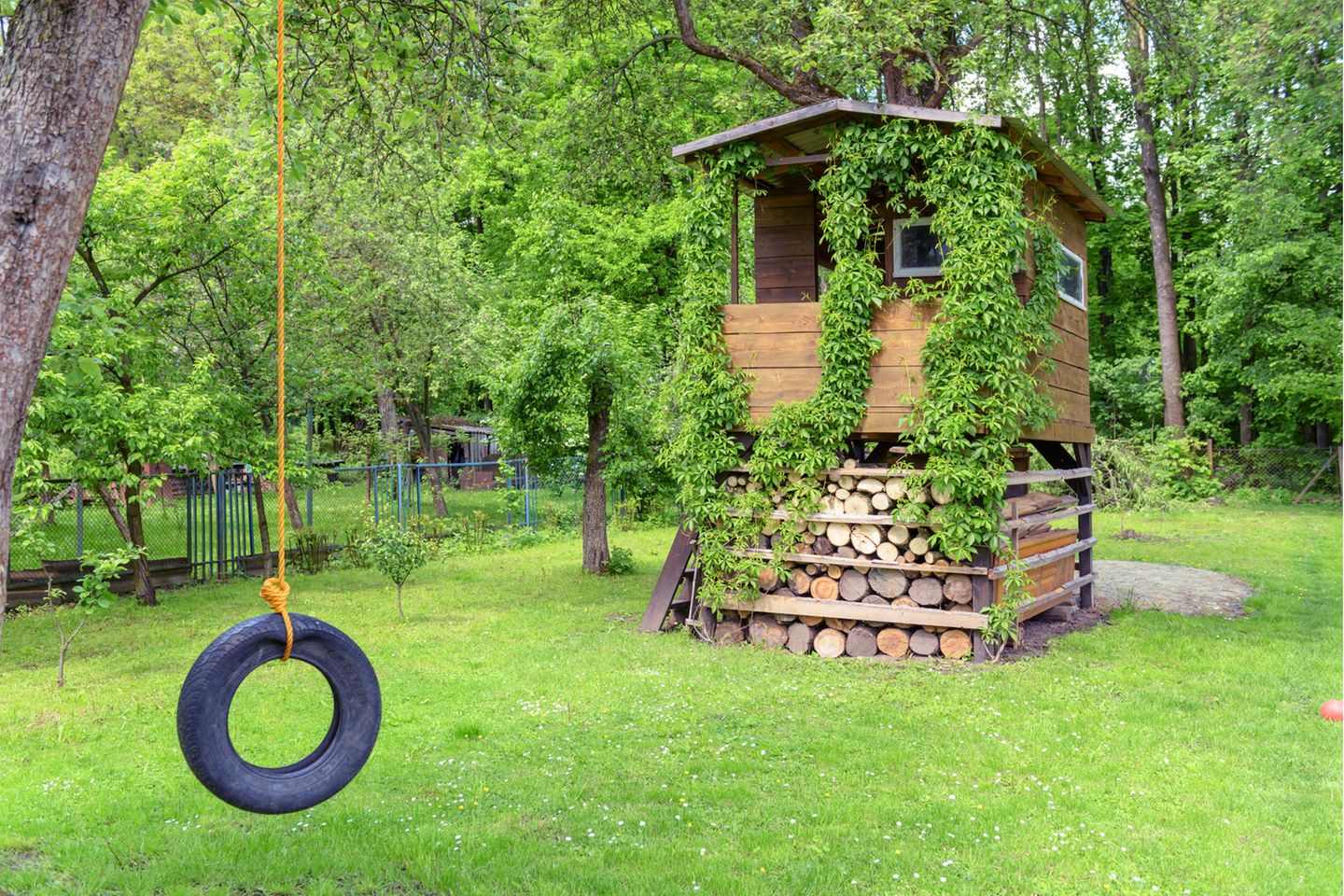 Spielhaus aus Holz, mit Kletterpflanzen bewachsen in einem baumbestandenen Garten