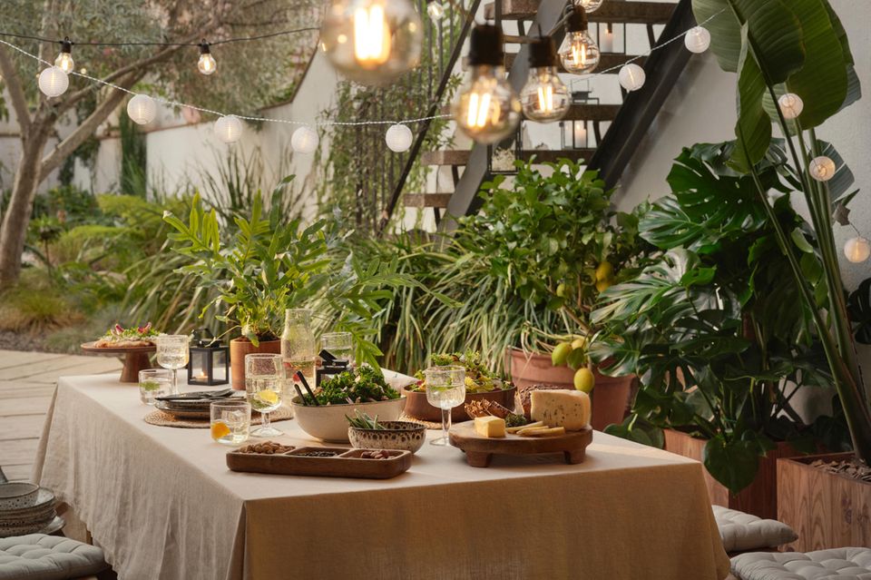 Ausladender Esstisch auf der Veranda mit Tischdecke, Geschirr, Topfpflanzen und Lichterketten