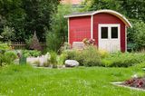 Gartenhaus mit halbovalem Dach in Schweden-Rot mit angelehnter Gartenbank vor einem Wiesenstück im Garten