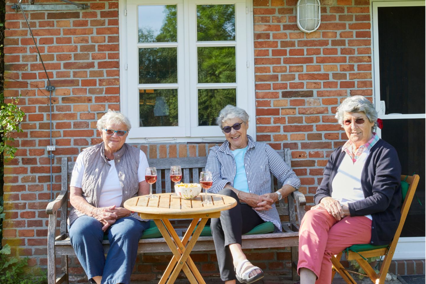 Drei ältere Damen mit Weingläsern vor reetgedecktem Haus