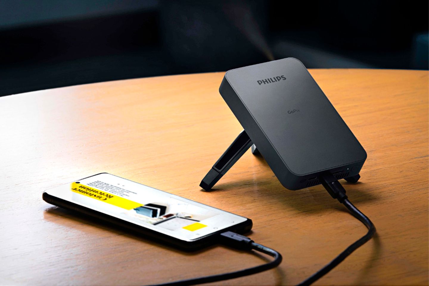 Kleiner schwarzer Projektor auf einem Holztisch, der via Kabel mit einen Smartphone verbunden ist