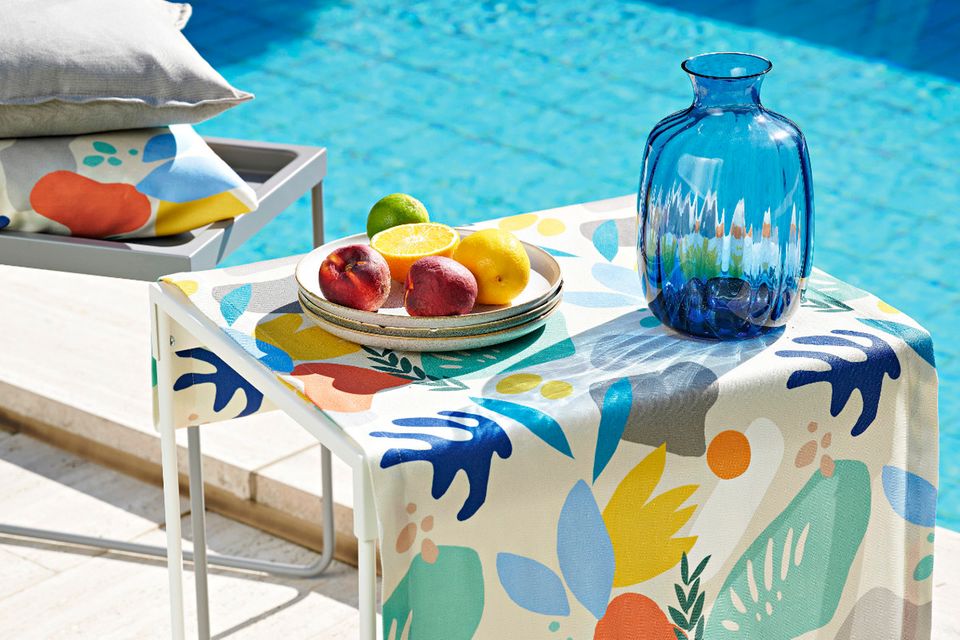 Simpler Gartentisch mit bunter Tischdecke, blauer Vase und Obst auf einem Teller vor einem Pool