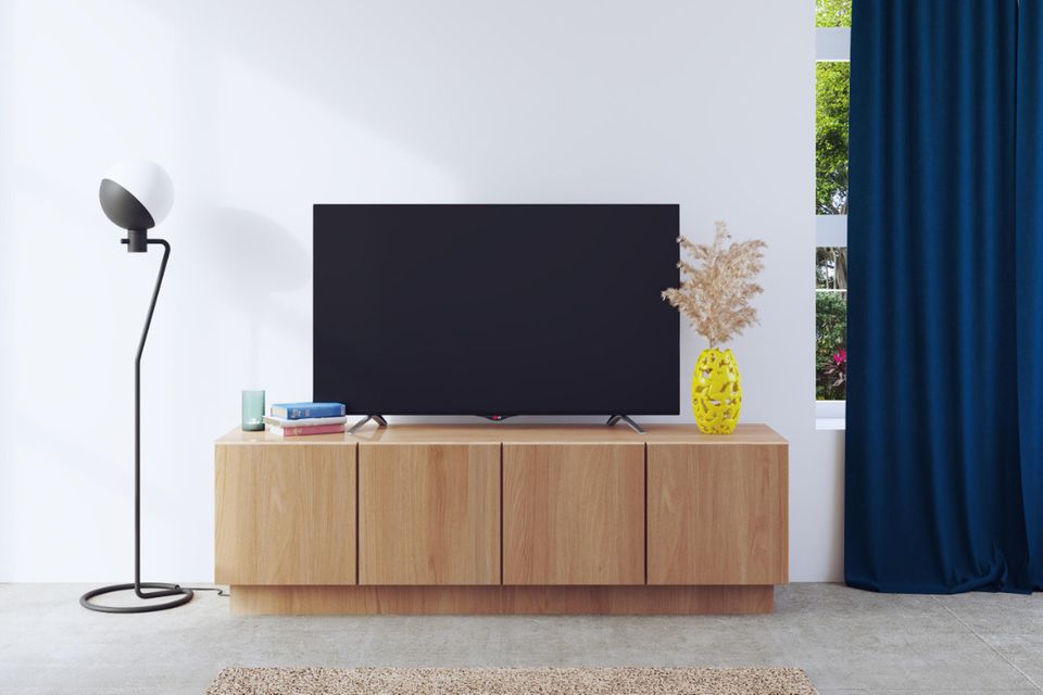 Fernsehtisch aus Naturholz vor einer weißen Wand mit Tischleuchte und blauen Vorhängen an den Seiten
