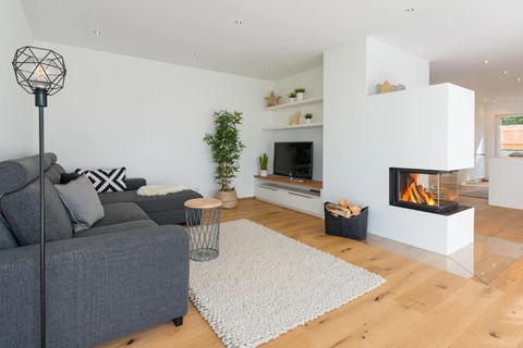 Wohnzimmer unter anderem mit grauer Sofalandschaft, weißem Hochflorteppich, Holzboden und Panoramakamin als Raumteiler