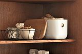 Körbe, Boxen und weiße Kisten auf einem Vorratsregal aus mittelbraunem Holz gefüllt mit Nüssen, Knoblauch und Müsli