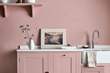 Rosafarbene Landhausküche vor einer rosafarbenen Wand; links ein Hocker und im Vordergrund ein grauweiß gefliester Boden