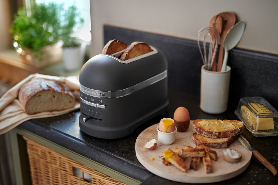 Dunkle Arbeitsplatte mit einem Toaster, einem Brot sowie einem Holzbrett mit getoasteten Butterbroten und zwei Eiern