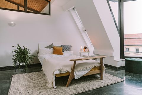 Bett im Dachgeschoss mit weißer Bettwäsche neben zwei großen Dachfenstern; auf dem Boden ein beiger Wollteppich