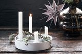 Adventsring aus Beton mit vier weißen Kerzen auf einem Holztisch; im Hintergrund ein Papierstern und eine Glasvase mit Zweig