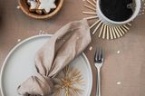 Beige eingedeckter Tisch im Topshot mit heller Keramik, Stoffserviette, Zimtsternen in einem Holzschüsselchen und Strohsternen