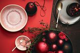Rosa gesprenkeltes Geschirr auf rubinrotem Tischläufer mit Gesteck aus granatapfelfarbenen Weihnachtskugeln