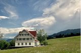 Kleines Hotel im Allgäu auf einem grünen Hügel vor einer Bergkulisse und blauem Himmel