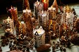 Zahlreiche Kerzenhalter aus Metall mit brennenden Kerzen auf dem Boden umgeben von Tannenzapfen und weiteren Naturmaterialien