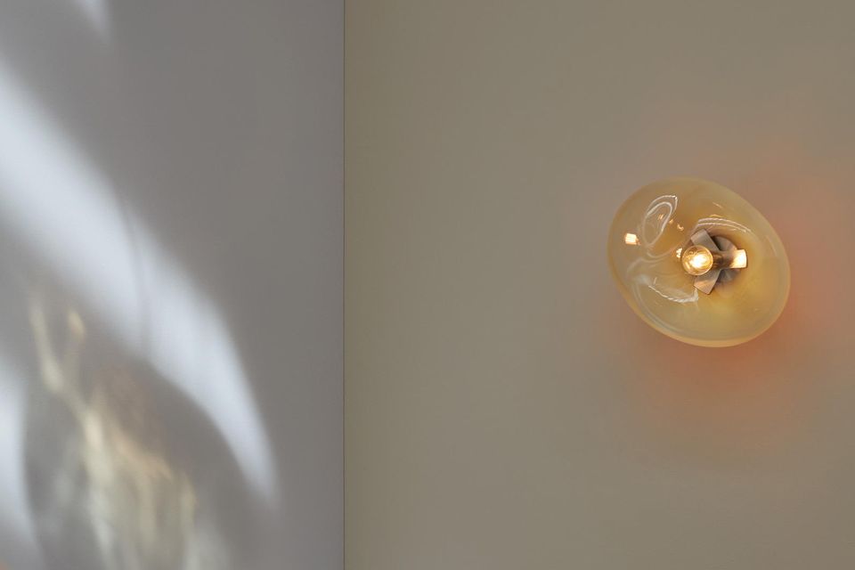 Organische Glasleuchte leuchtet weiches Licht, während Tageslicht durch sie hindurchfällt und Lichtspiele auf eine Wand wirft