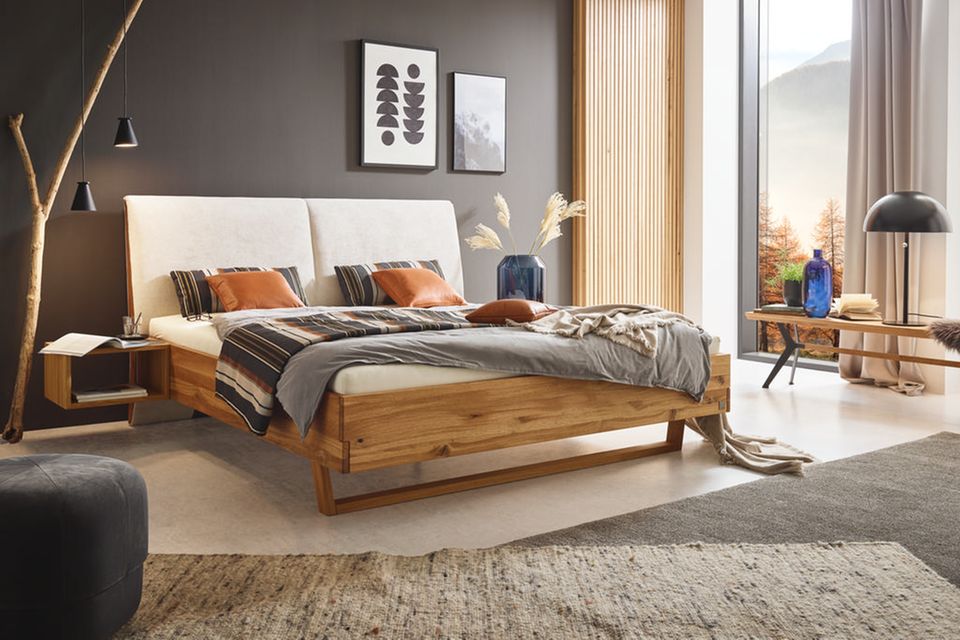 Massivholzbett mit cremefarbenem Bezug in einem Schlafzimmer mit grauer Wand und bodentiefen Fenstern