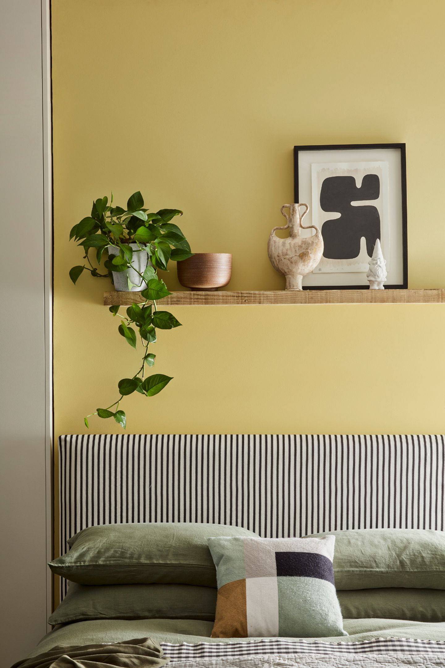Gelbe Wand mit Regalbrett mit Deko und Grünpflanze hinter Betthaupt