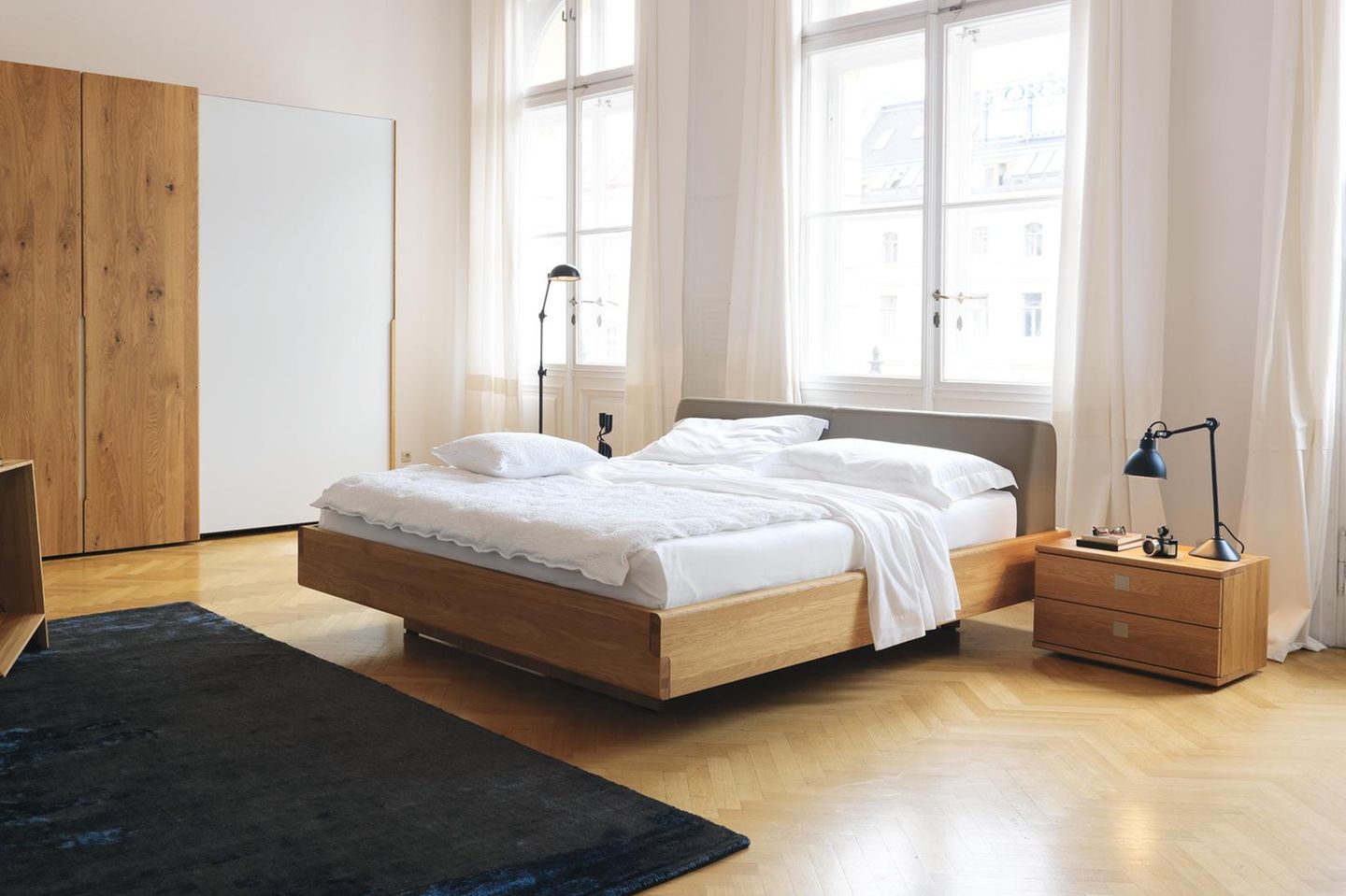 Ein helles Holzbett mit grauem Lederheadboard steht in einem Raum mit Fischgrätparkett, Holznachtisch und Holzkleiderschrank