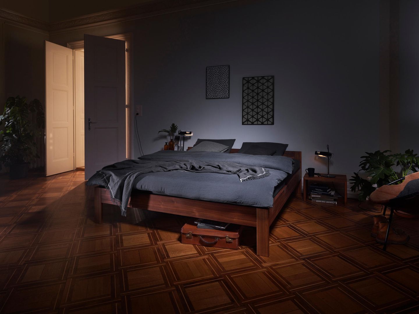 Ein dunkles Holzbett mit grauer Bettwäsche steht in einem abgedunkelten Raum