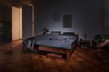 Ein dunkles Holzbett mit grauer Bettwäsche steht in einem abgedunkelten Raum