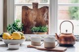 Ein Kupferkessel, zwei Keramikbecher und eine Schale Zitronen vor einem Altbaufenster mit Sprossen.