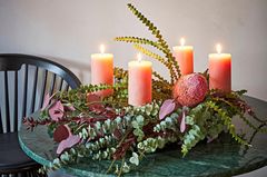 Adventskranz mit lachsfarbenen Kerzen, Proteablüten und Eukalyptus auf einem schwarzen Tisch.
