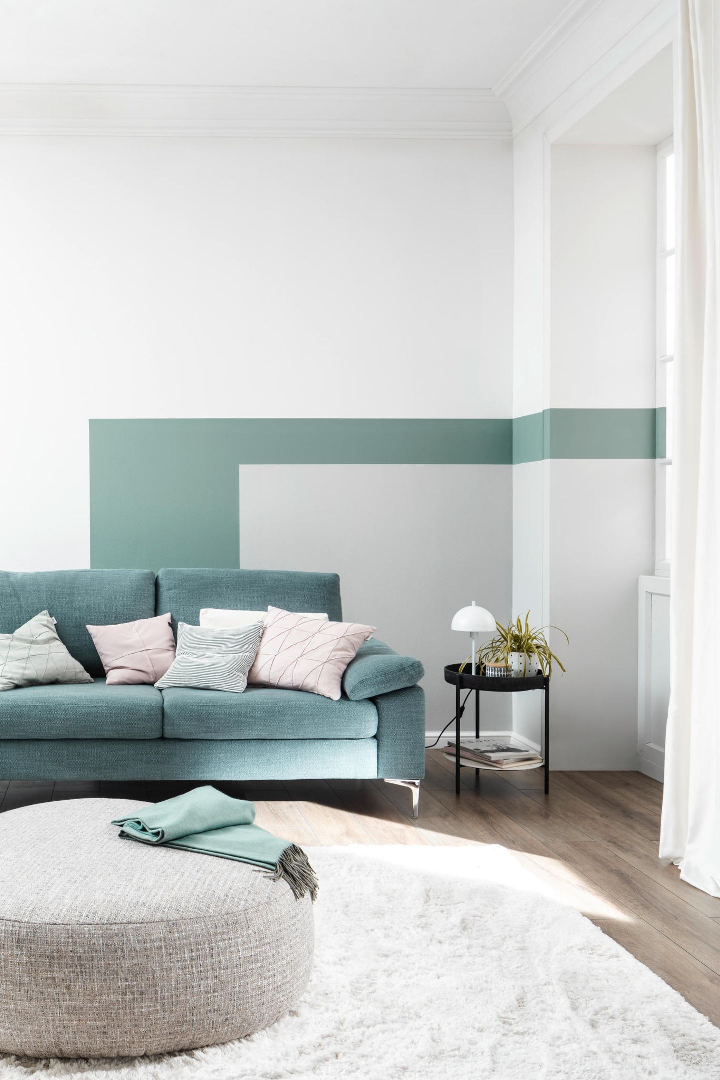 Wohnzimmer mit blaugrünem Sofa und grauem Pouf vor einer farbigen Wand in Grün-Grau