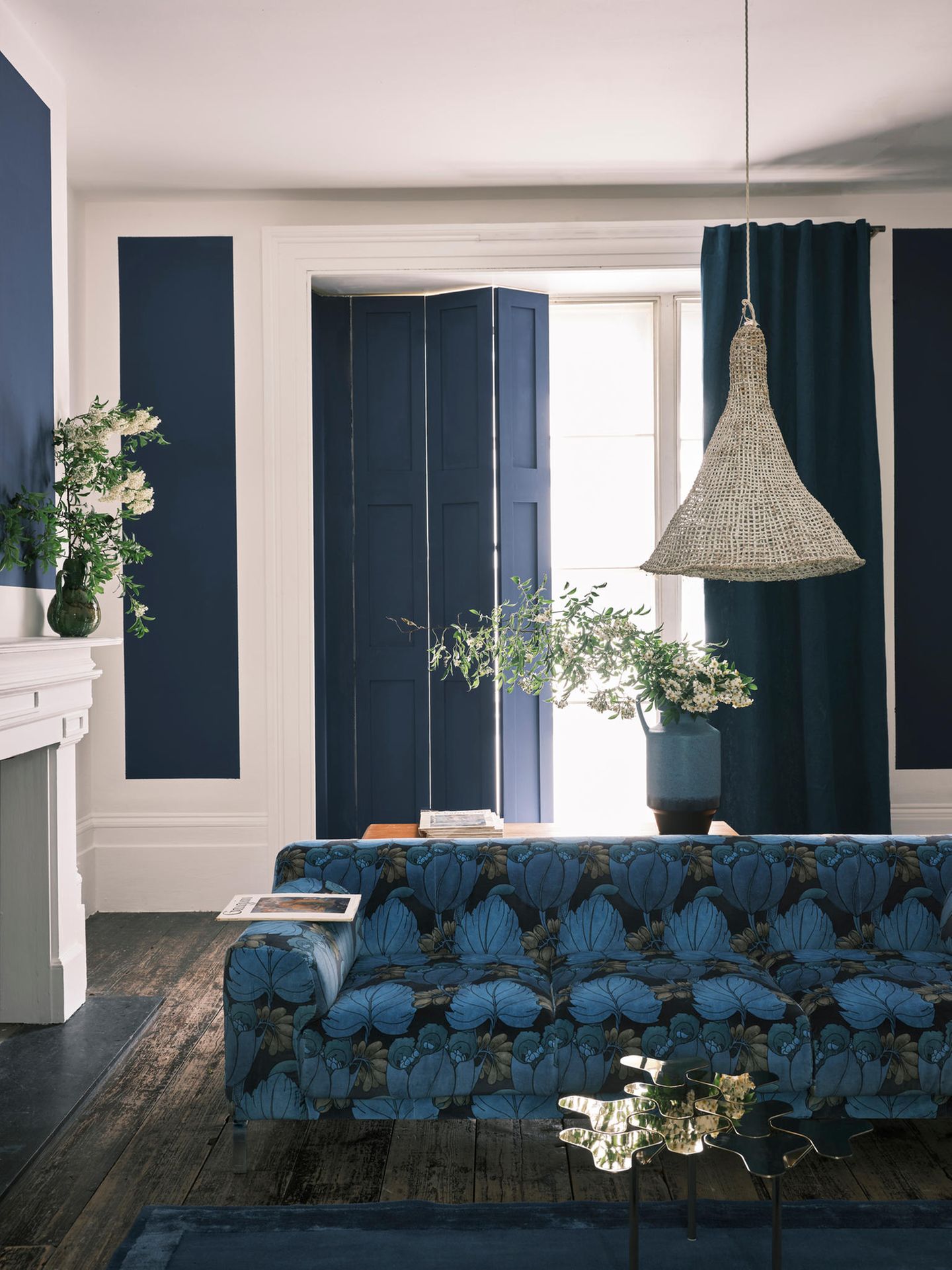 Wohnzimmer mit weißen Wänden und blauen Akzenten sowie blau-schwarzem Mustersofa und einem weiß lackierten Kaminsims