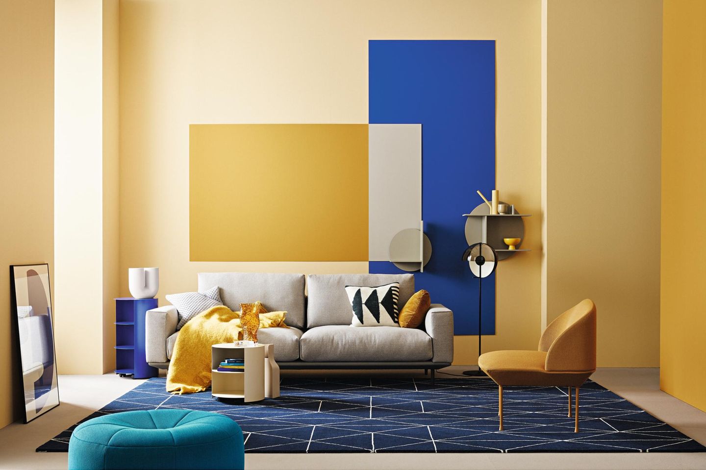 Wohnzimmer mit gelb-blau gestrichenen Wänden grauem Sofa und petrolfarbenem Pouf