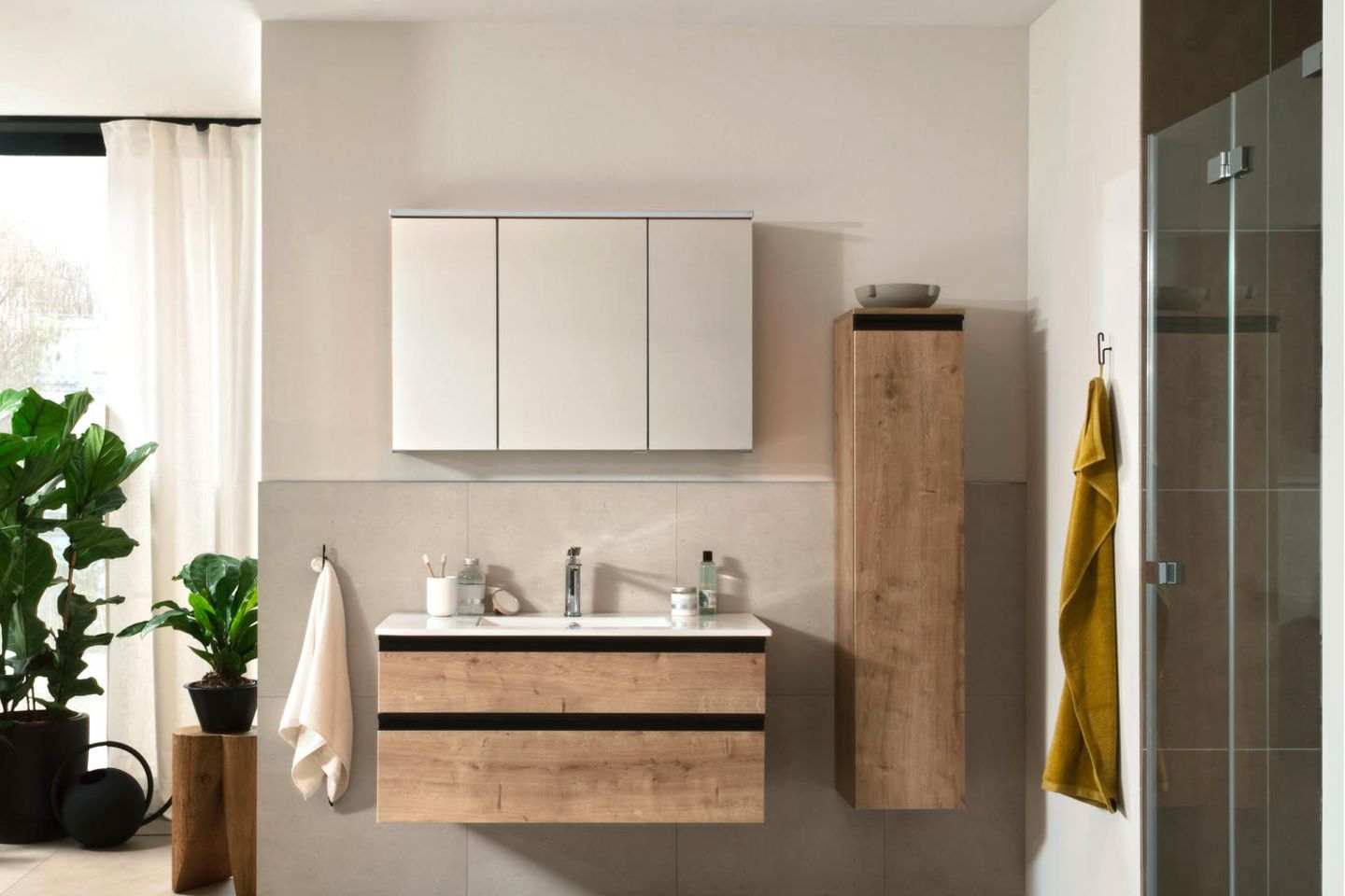 Badezimmer mit Möbel in Holzoptik, einem Waschtisch, Hängeschrank, Dusche und verschiedenen Grünpflanzen.