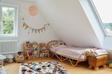 Kinderzimmer mit Korbmöbeln, Spielsachen und einem Kinderbett mit rosafarbener Tagesdecke.