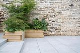 Mit natürlichen Gehwegplatten gepflastertes Hanggrundstück mit Grünpflanzen in bepflanzten Hochbeeten vor alter Steinmauer