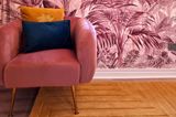 Palmentapete in Pink mit einem pinkfarbenem Sessel und Kissen in Blau und Gelb.