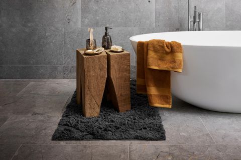 Neben einer freistehenden Badewanne steht ein Holzblock als Beistelltischchen; auf den grauen Steinfliesen liegt ein Teppich