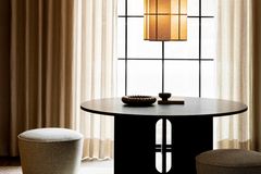 Dunkler Esszimmertisch, zwei Poufs sowie eine Pendelleuchte vor einem hellen Fenster mit Gardinen und Vorhängen