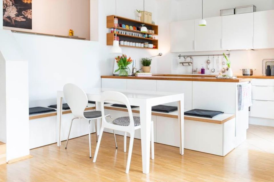 Weiße Einbauküche mit Arbeitsplatten aus Holz und einem weißen Esstisch mit Sitzbank im Vordergrund
