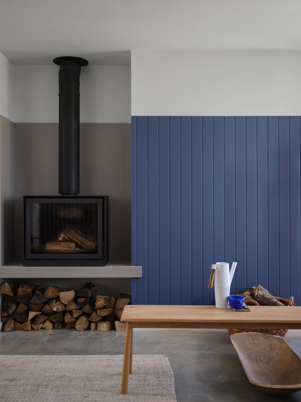 Blaue Wandvertäfelung mit breitem weißen Rand und schwarzem Ofen, rechts eine Holzbank mit weißer Gießkanne