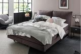 Anthrazitfarbenes Bett mit Decken und Kissen in Grautönen vor großer Fensterfront