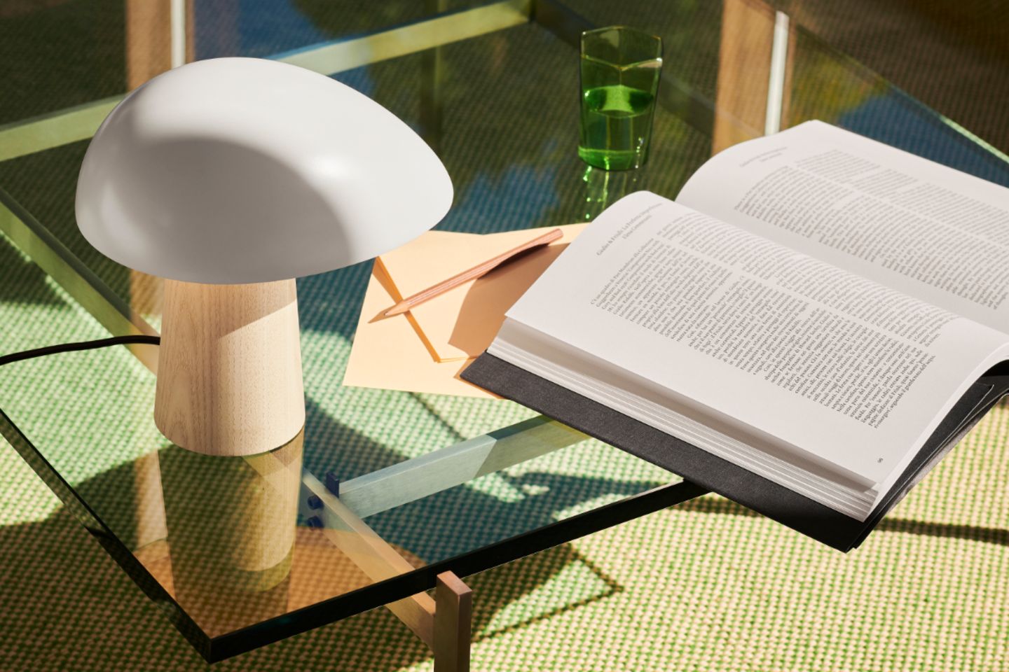 Tischleuchte aus mit Holzsockel und weißem Schirm auf Beistelltisch aus Glas mit Buch und Notizzetteln