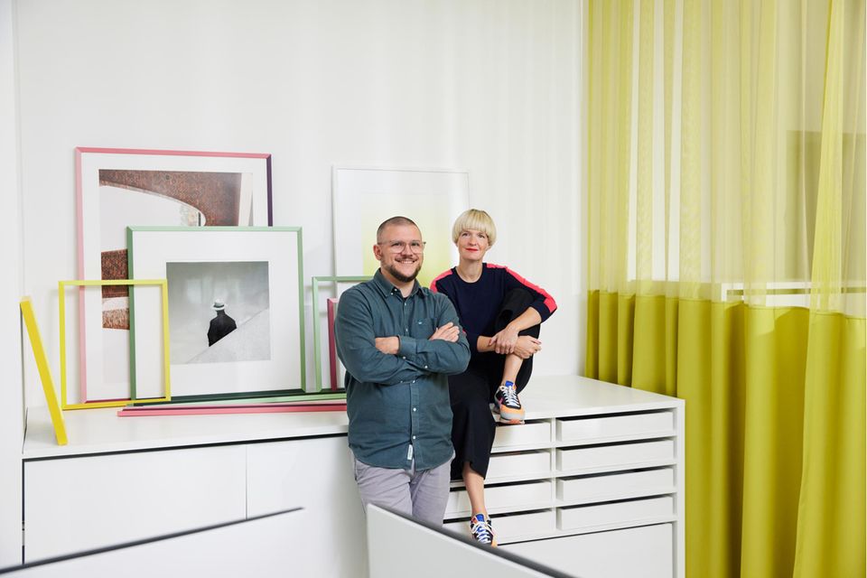 Mann und Frau vor weißem Sideboard mit bunten Bilderrahmen im Hintergrund