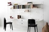 Eine weiße Bücherregal-Schreibtisch-Kombination steht vor einer weißen Wand mit schwarzen Stühlen.