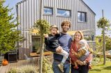 Familie mit zwei Kindern im Garten vor grauem Holzhaus